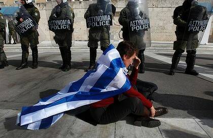 An anti-austerity protester draped with a Greek flag sits in front of police guarding parliament in Athens.
